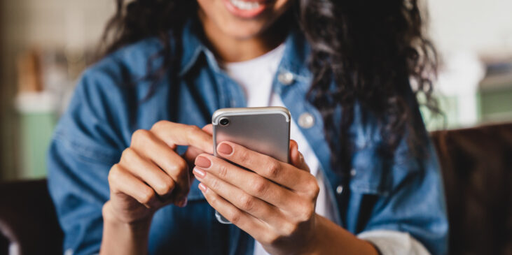 Cropped shot of a young woman using smart phone at home. Smiling and using smartphone at home, messaging or browsing social networks while relaxing on couch