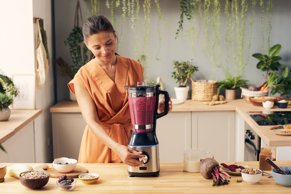 Image of a woman pressing start button of blender during preparation of homemade vegetable smoothie by kitchen table